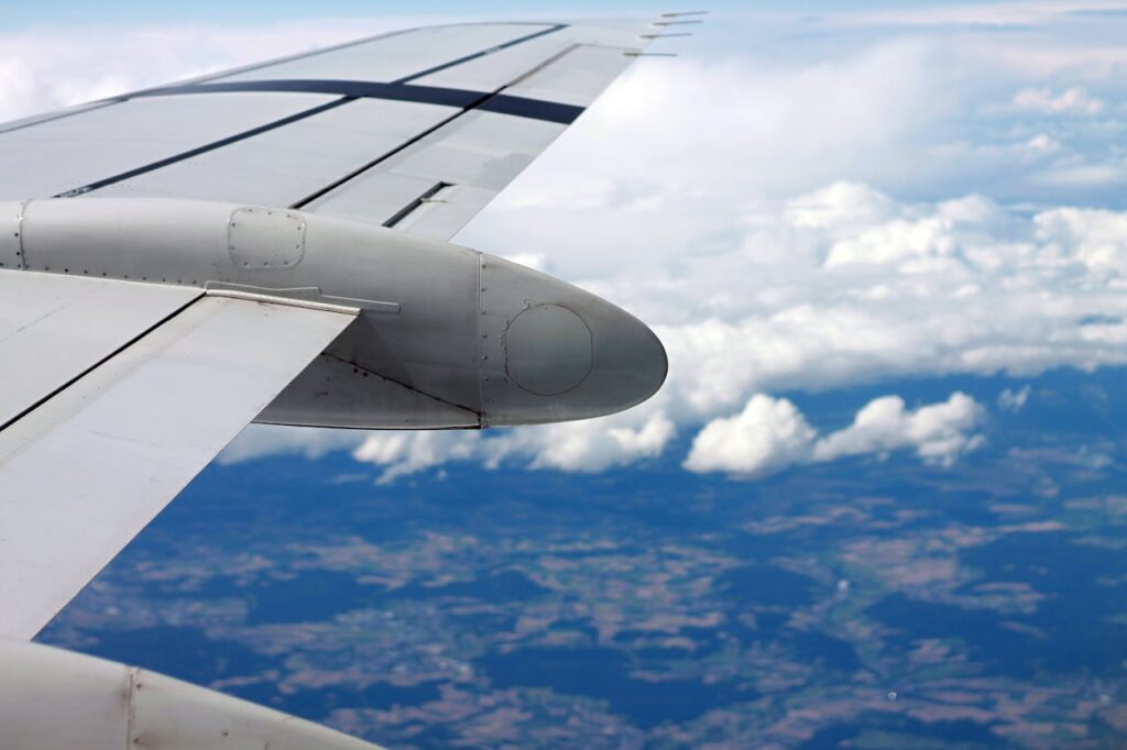 View of an airplane wing flying over clouds and landscape below, showcasing aerial travel.